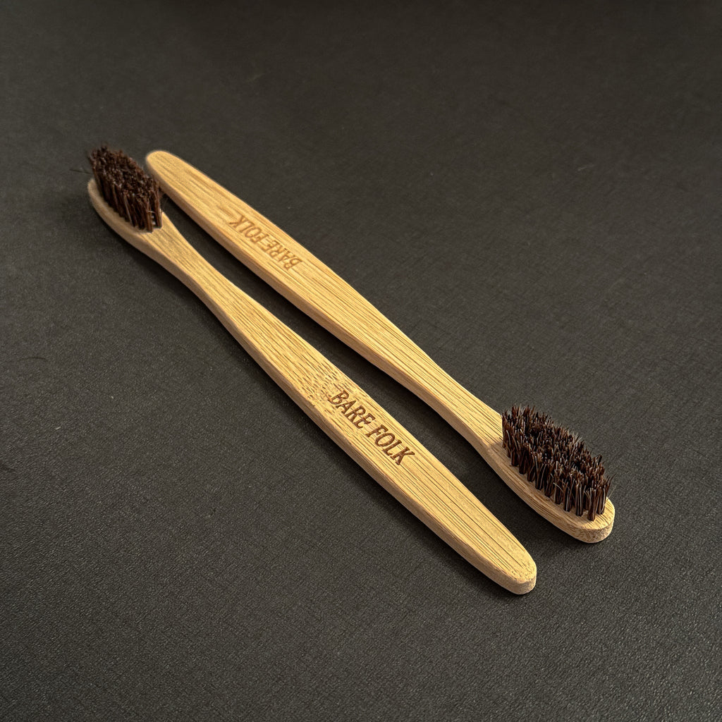 Two bamboo toothbrushes with dark brown horse bristles, one engraved with 'BARE FOLK' and the other with 'BARE FOLK', displayed diagonally on a dark textured background.
